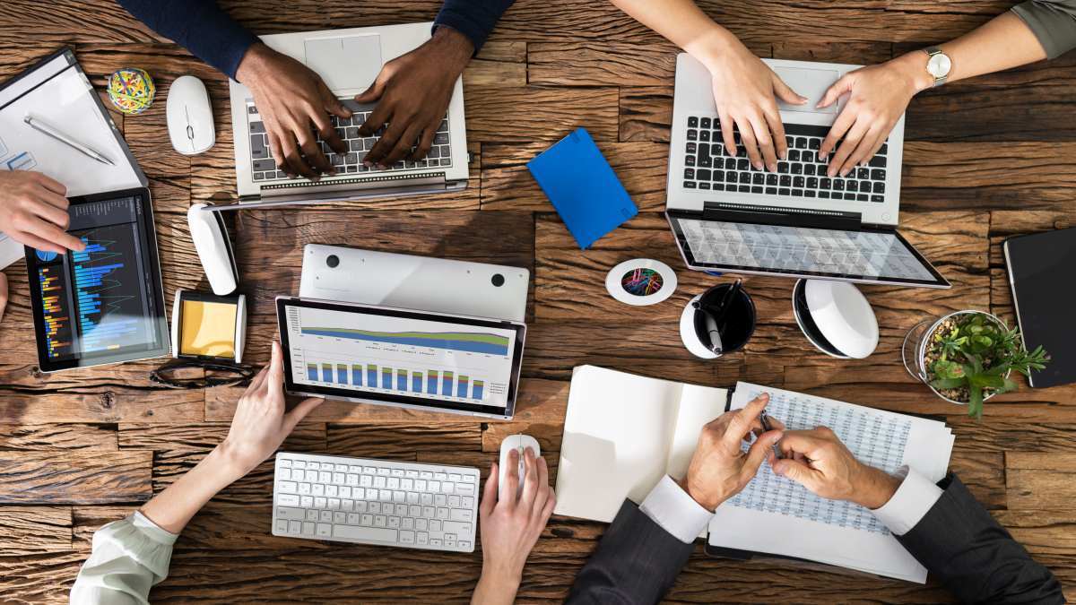 mehrere-personen-arbeiten-an-laptops-und-tablets-auf-einem-holztisch-einige-tippen-und-andere-analysieren-diagramme mehrere-personen-arbeiten-an-laptops-und-tablets-auf-einem-holztisch-einige-tippen-und-andere-analysieren-diagramme